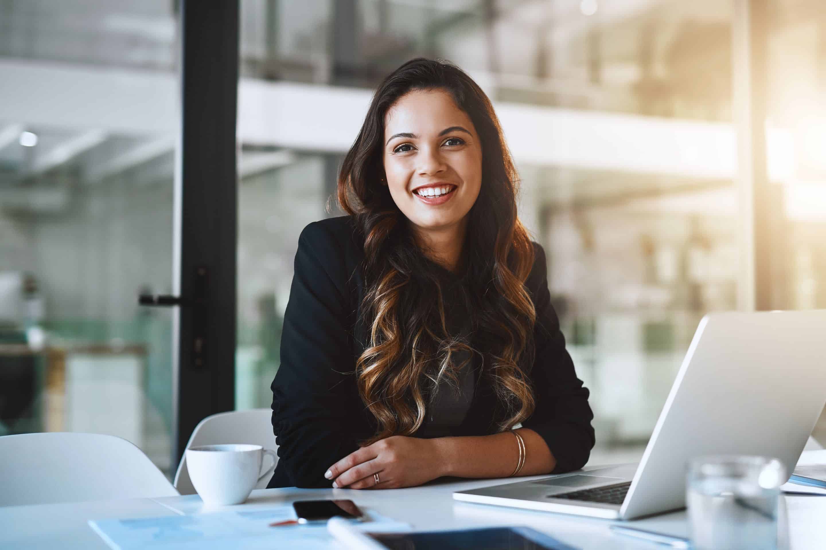 Evaluate Your Career Progression - Young woman in the office sitting at her desk and smiling in front of her computer